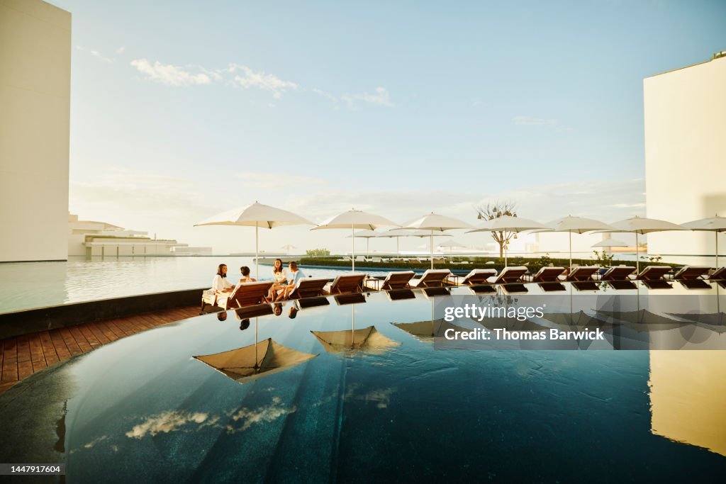 Wide shot of family hanging out on lounge chairs by pool at resort