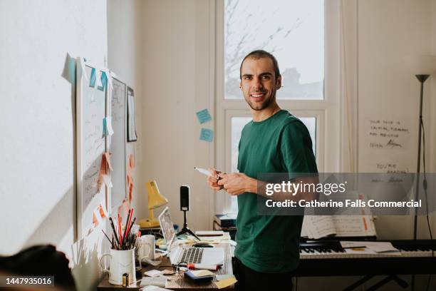 a man in a small home-office stands by a disorganised desk, holding a white board marker, and smiles - man made stock pictures, royalty-free photos & images