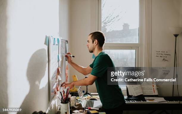 a man leans over a messy desk and writes on a wall-mounted whiteboard in a home-office environment - writing list stock pictures, royalty-free photos & images