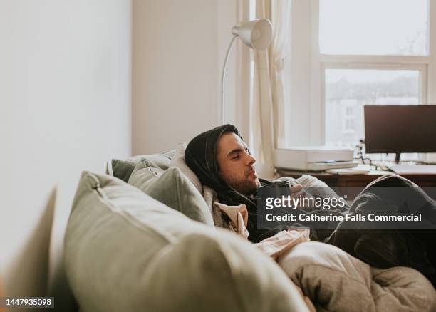 a relaxed man in a bathrobe reclines on a soft couch, surrounded by a duvet, and pillows. he enjoys a moment of peace with a hot drink. - couch potato stock-fotos und bilder