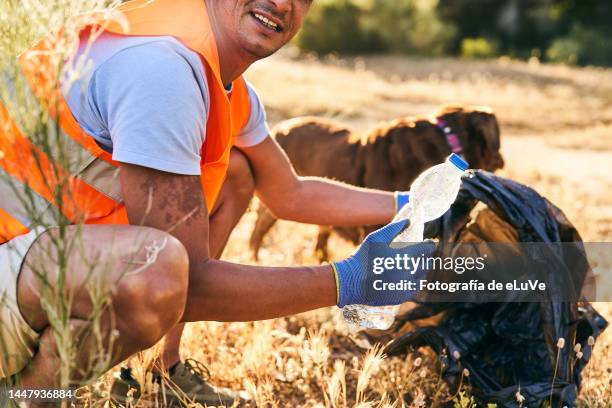 man crouching pick up plastic bottle to be dumped in the trash looking at camera - orange glove stock pictures, royalty-free photos & images