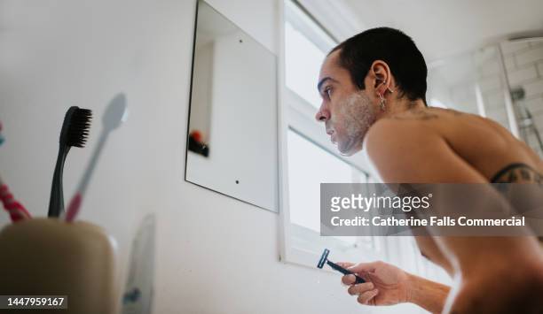 a man looks into a bathroom mirror and shaves with a manual razor - man shaving foam stock pictures, royalty-free photos & images