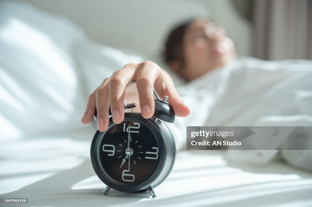 Young asian woman wake up early in morning. Female lying on bed and trying turn off alarm clock. Young sleeping woman and alarm clock in bedroom at home.
