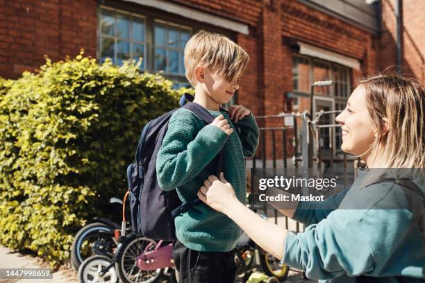 happy mother looking at son with backpack outside school building - back to school stock pictures, royalty-free photos & images