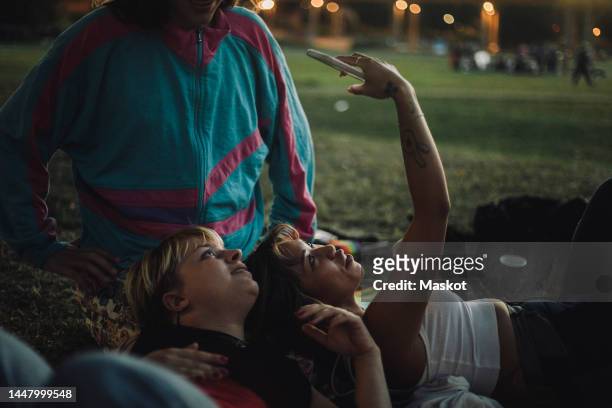 young woman taking selfie with non-binary person while lying on friend's lap during sunset - stockholm park stock pictures, royalty-free photos & images