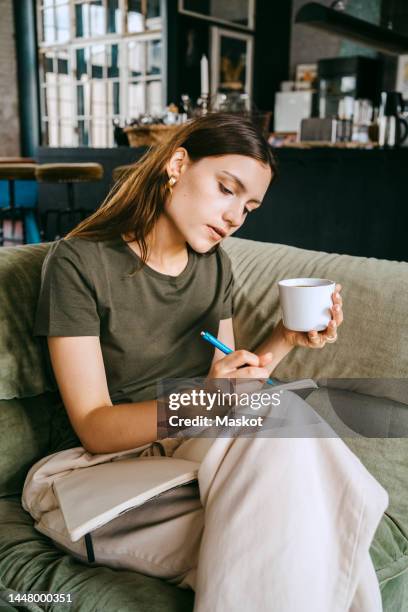 young female entrepreneur writing in book holding coffee cup while sitting on sofa in studio - dreiviertelansicht stock-fotos und bilder