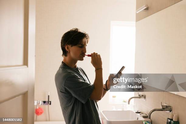 side view of young man using smart phone while brushing teeth in bathroom at home - brushing teeth stock pictures, royalty-free photos & images