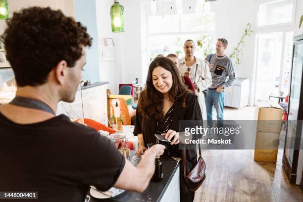 young female customer paying through tap and pay while standing in queue at cafe - customers lining up stock pictures, royalty-free photos & images