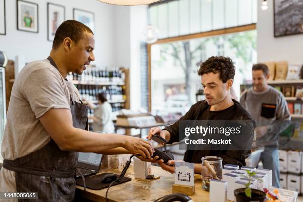 side view of male owner assisting customer doing online payment at checkout counter in store - apple pay stock pictures, royalty-free photos & images