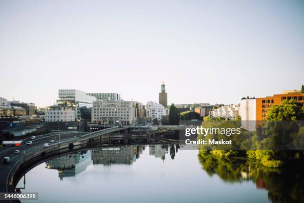 kungsholmen town hall amidst modern buildings reflected in sea against clear sky - stockholm buildings stock pictures, royalty-free photos & images