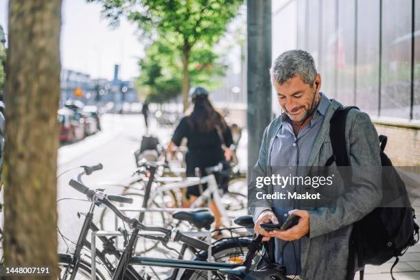 smiling mature businessman using smart phone while standing at bicycle parking station - bicycle parking station stock pictures, royalty-free photos & images