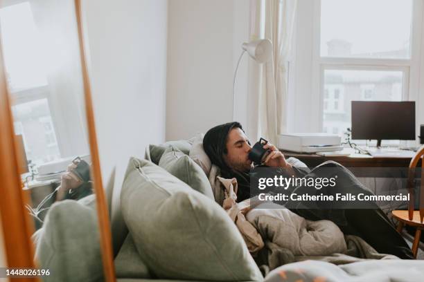 a man reclines on a soft couch, in a bath robe. he sips on a hot drink. - hangover stock pictures, royalty-free photos & images