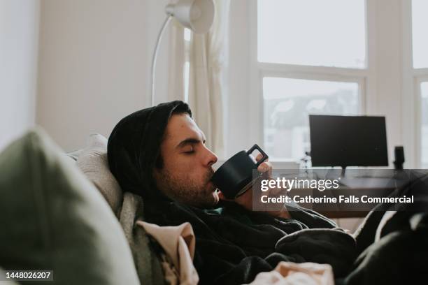 a man reclines on a soft couch, in a bath robe. he sips on a hot drink. - taking a bath stock-fotos und bilder