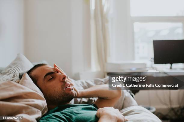a man wakes up on a sofa, surrounded by sheets and duvets and pillows. he rubs his eyes and face as he tries to rouse himself. - yawning is contagious imagens e fotografias de stock