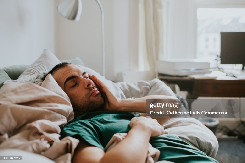 A man wakes up on a sofa, surrounded by sheets and duvets and pillows. He rubs his eyes and face as he tries to rouse himself.