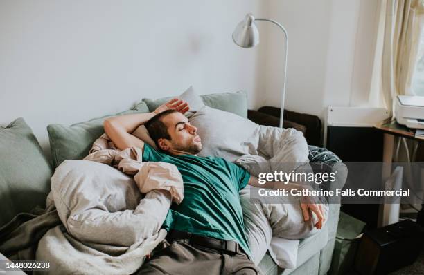 a man is asleep on a sofa surrounded by duvets and pillows - luiheid stockfoto's en -beelden
