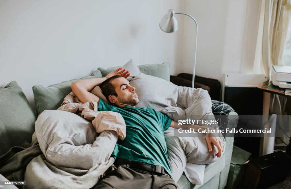 A man is asleep on a sofa surrounded by duvets and pillows