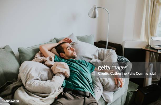 a man is asleep on a sofa surrounded by duvets and pillows - procrastination stock pictures, royalty-free photos & images