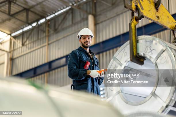 warehouse and storage management in a coil steel manufacturing industry. a male overhead crane operator hooks lifting rolled-up metal sheets in a coil sheet metal warehouse. safety culture in manufacturing industry. - metalwork stock-fotos und bilder
