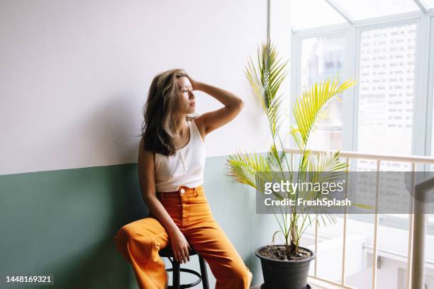 uma mulher bonita e feliz em camiseta branca e calças laranjas olhando para o lado enquanto relaxa no corredor de seu prédio - calças verdes - fotografias e filmes do acervo