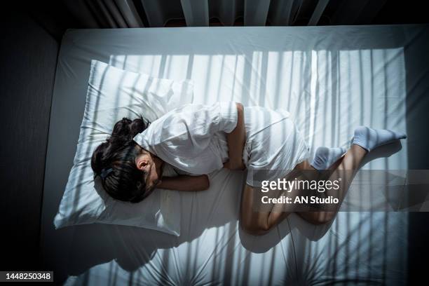 young woman suffering from abdominal pain while lying on bed at home. sick young asian woman lying in white bed. holding her belly. top view - bauchweh stock-fotos und bilder