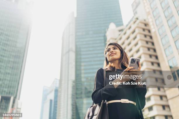 confident young businesswoman with smartphone standing against modern buildings - fato completo imagens e fotografias de stock