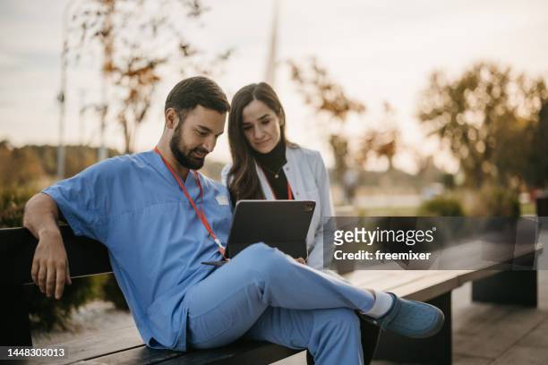 male and female doctors sitting on a bench in front of hospital and using digital tablet - handsome doctors stock pictures, royalty-free photos & images
