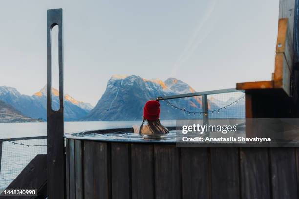 rear view of a woman in red hat relaxing in hot tub with scenic view of the winter fjord in norway - norway winter stock pictures, royalty-free photos & images