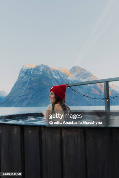 vista laterale di una donna in cappello rosso che si rilassa nella vasca idromassaggio con vista panoramica del fiordo invernale in norvegia - sauna winter foto e immagini stock