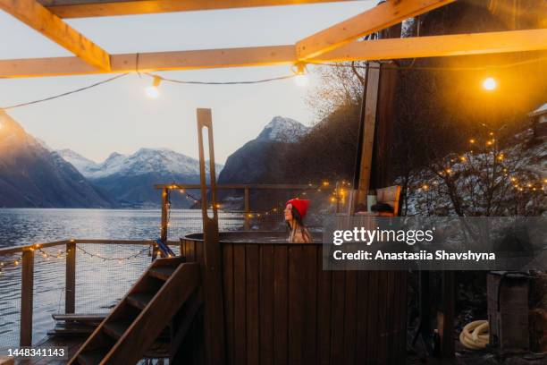 seitenansicht einer frau mit rotem hut, die sich im whirlpool entspannt, mit malerischem blick auf den winterfjord in norwegen - sauna winter weihnachten stock-fotos und bilder
