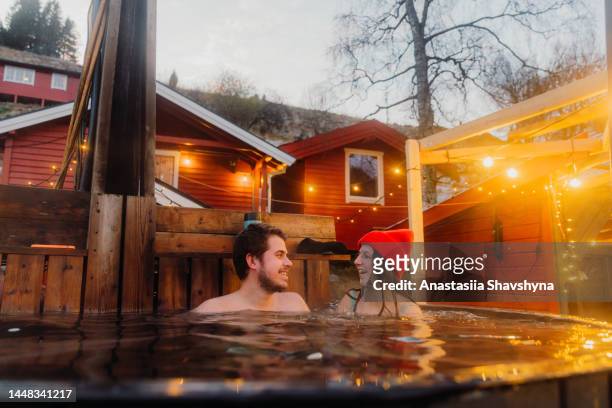 happy woman and man relaxing in hot tub with scenic mountain view by the fjord in winter in norway - bad weather stockfoto's en -beelden