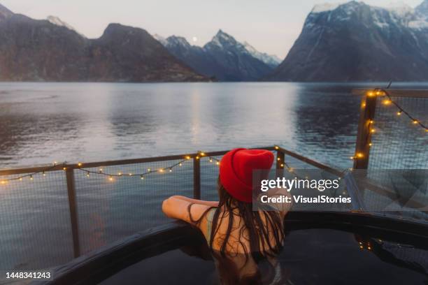 rear view of a woman in red hat relaxing in hot tub with scenic view of the winter fjord in norway - hygge stockfoto's en -beelden