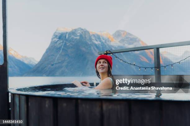 seitenansicht einer glücklichen frau mit rotem hut, die sich im whirlpool mit malerischem blick auf den winterfjord in norwegen entspannt - sauna stock-fotos und bilder