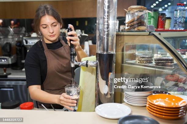 a young beautiful woman in a brown apron is standing and pouring water into a glass. the concept of a small business. work for yourself. - part time worker fotografías e imágenes de stock