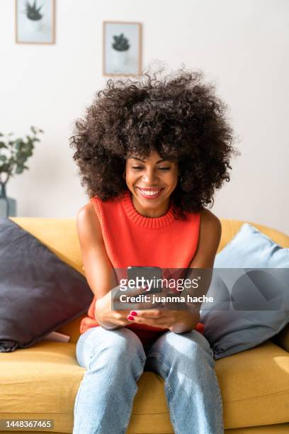 smiling woman sitting in living room while texting with mobile phone to her friends to have lunch together. . - african lady sitting and looking at her smartphone stock-fotos und bilder