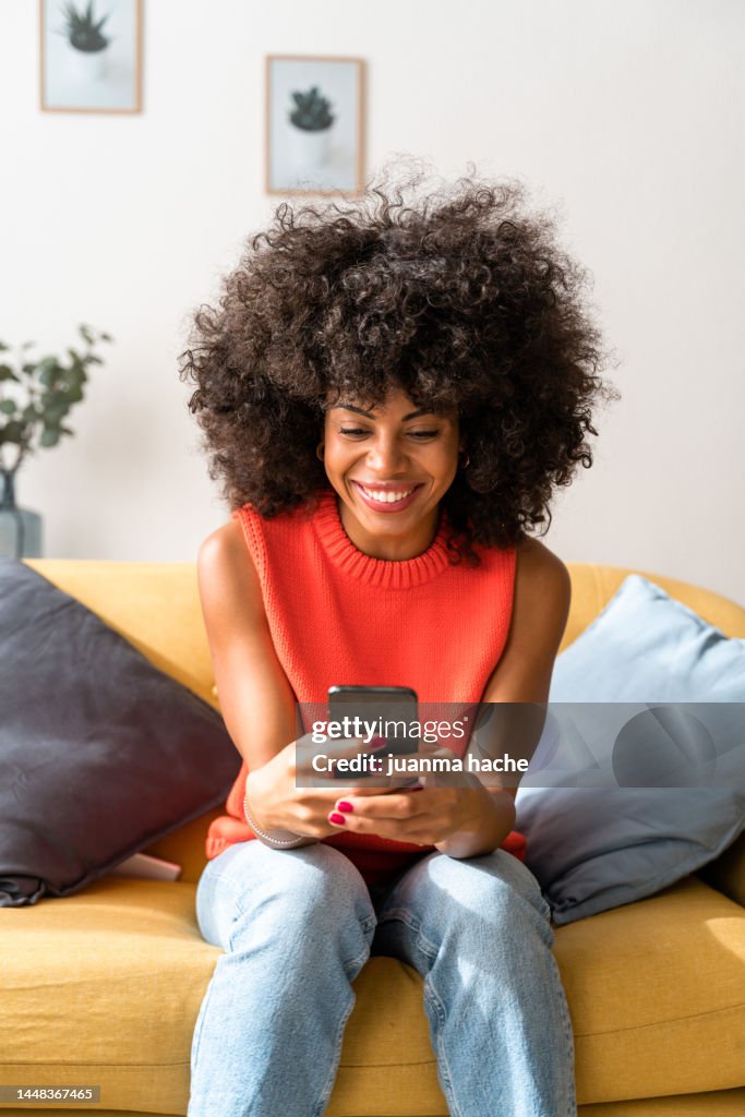Smiling woman sitting in living room while texting with mobile phone to her friends to have lunch together. .