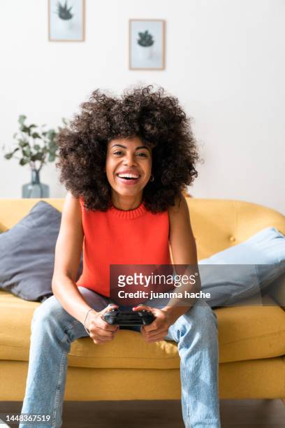 optimistic woman sitting on living room sofa holding video game controller. - manette photos et images de collection