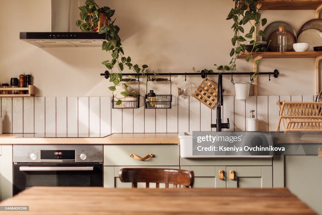 Empty kitchen island with marble surface in foreground, green vintage countertop with drawers and pendant lights hanging above, lots of flowers in jars, blurred background