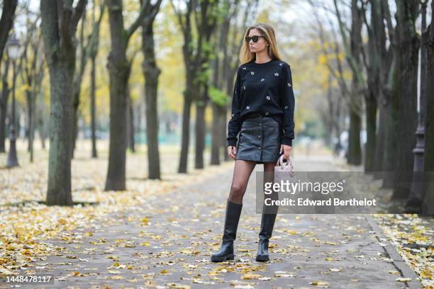 Mathilde Frachon wears black sunglasses, gold earrings, a black with embroidered white and pale pink bees pattern pullover, a black shiny leather...