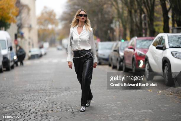 Natalia Verza wears black squared sunglasses from Prada, silver earrings, a white silk buttoned shirt, a silver shiny varnished leather shoulder bag,...