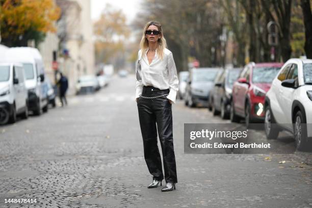 Natalia Verza wears black squared sunglasses from Prada, silver earrings, a white silk buttoned shirt, a silver shiny varnished leather shoulder bag,...