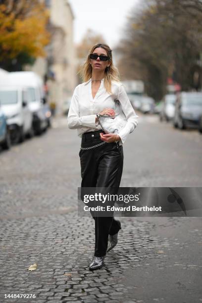 Natalia Verza wears black squared sunglasses from Prada, silver earrings, a white silk buttoned shirt, a silver shiny varnished leather shoulder bag,...