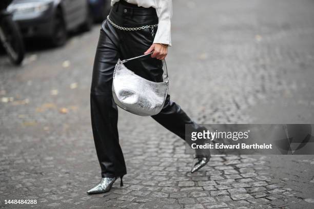 Natalia Verza wears a white silk buttoned shirt, a silver shiny varnished leather shoulder bag, black shiny leather large wide legs pants, a silver...