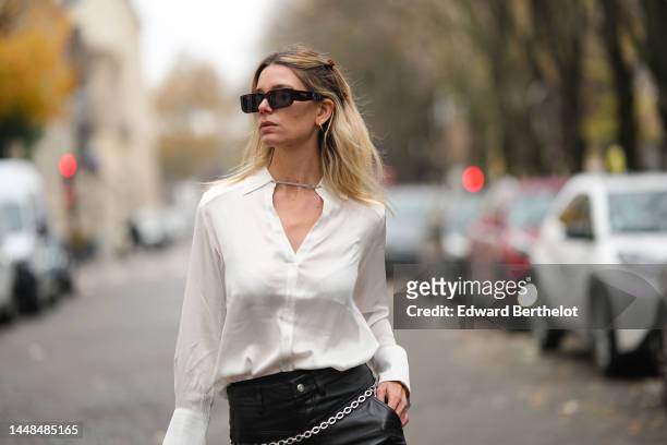 Natalia Verza wears black squared sunglasses from Prada, silver earrings, a white silk buttoned shirt, black shiny leather large wide legs pants, a...