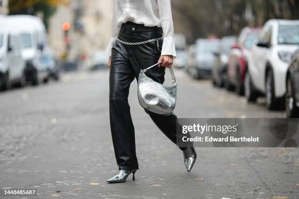 Natalia Verza wears a white silk buttoned shirt, a silver shiny varnished leather shoulder bag, black shiny leather large wide legs pants, a silver...