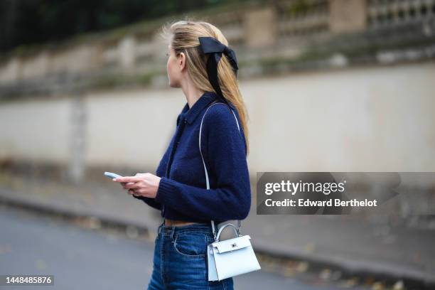 Mathilde Frachon wears a black ribbon knot in the hair, a navy blue wool / crystals buttons / cropped pullover, a blue white shiny leather mini Kelly...