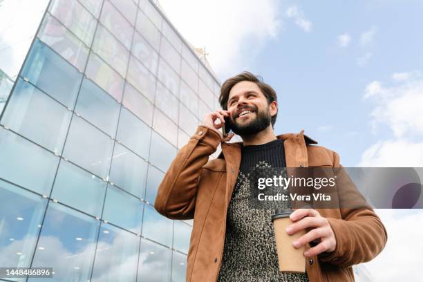 young man with beard and brown clothes using technology outdoors holding coffee - warm jacket stock pictures, royalty-free photos & images