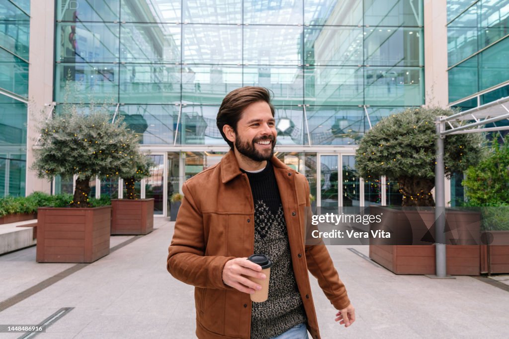 Young latin american man with beard and brown jacket walking in the street holding coffee