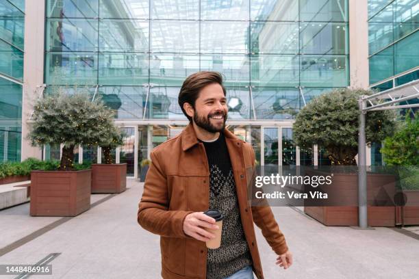 young latin american man with beard and brown jacket walking in the street holding coffee - takeaway coffee cup stock pictures, royalty-free photos & images