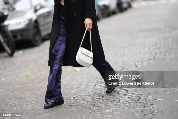 Mathilde Frachon wears a black long buttoned coat, a white shiny leather Cleo shoulder bag from Prada, purple satin silk large flared pants, gray...